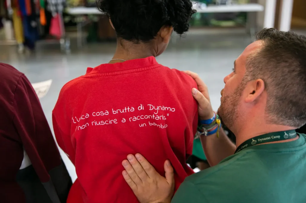 Operatore Dynamo Camp con una mano sulla spalla di un bambino che indossa una maglietta con la scritta: “L’unica cosa brutta di Dynamo è non riuscire a raccontarlo.”